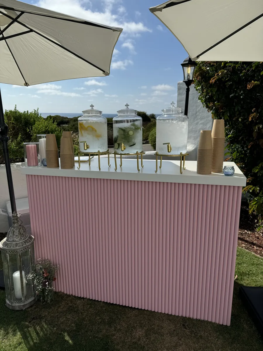 A pink fluted bar on a lawn by the coast, with drink dispensers, cups and two white umbrellas